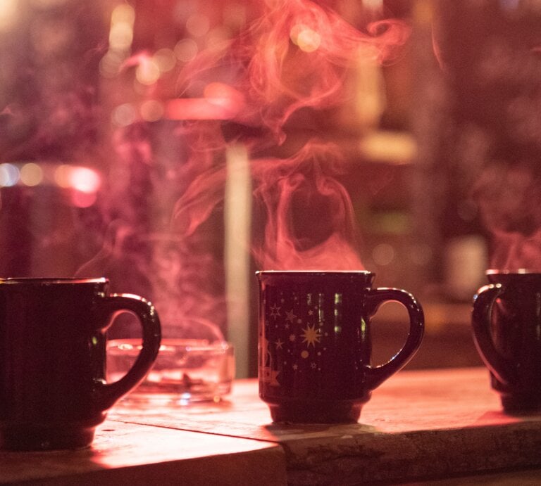 Drei dampfende Tassen Glühwein auf einem Holztisch, unscharfer Weihnachtsmarkt im Hintergrund mit Lichterglanz. | © Adobe Stock/ Marco Martins