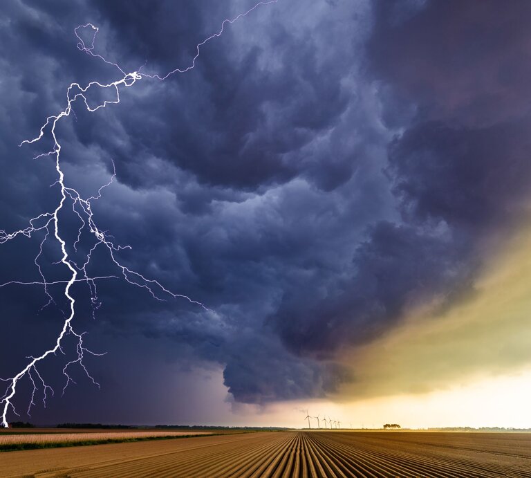 Ein Blitz erhellt dramatisch den dunklen Himmel über einem abgeernteten Feld mit Windrädern im Hintergrund. | © GettyImages/George Pachantouris