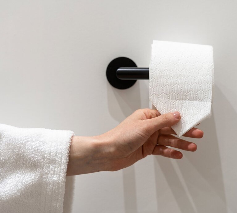 Eine Hand im weißen Bademantel greift nach einem dreieckig gefalteten Blatt Toilettenpapier an einer schwarzen Wandhalterung. | © Adobe Stock/ brizmaker