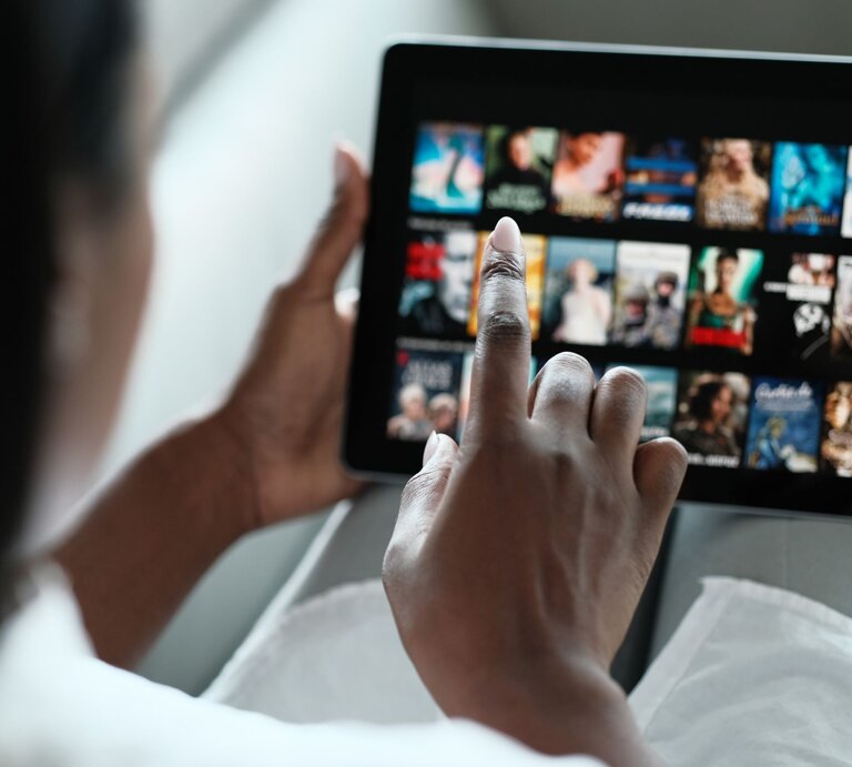 Eine Person mit dunkler Hautfarbe wählt auf einem Tablet im Querformat einen Film aus einer Streaming-Bibliothek aus. | © Adobe Stock/ Diego Cervo