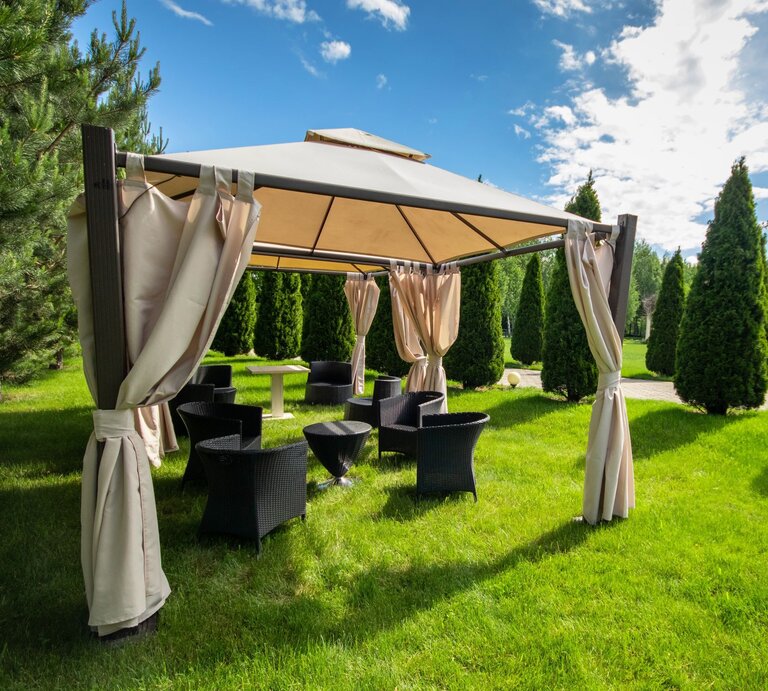Ein luftiger Gartenpavillon mit offenen Vorhängen lädt inmitten symmetrisch gepflanzter Koniferen zum Verweilen ein. | © Adobe Stock/ Roman