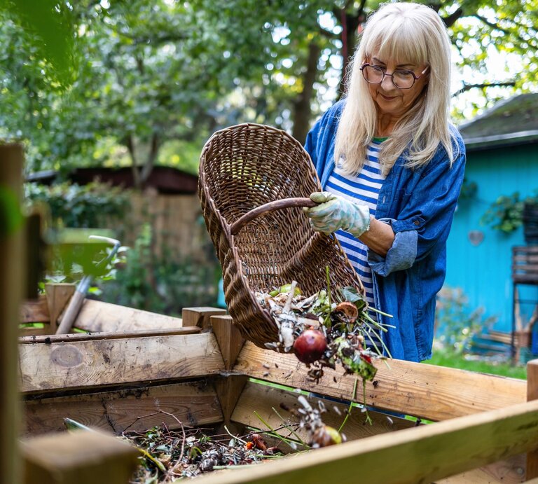 Eine ältere Frau mit blondem Haar und Brille leert lächelnd einen Korb mit Gartenabfällen in einen Kompostbehälter in grünem Garten. | © Shutterstock/pikselstock