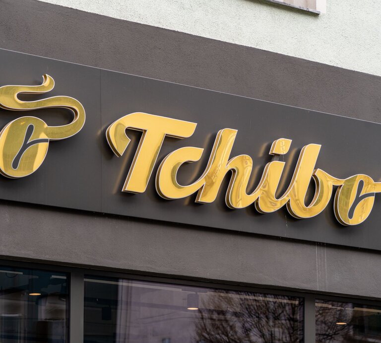 Goldenes Tchibo-Logo auf schwarzem Hintergrund an einer Hausfassade, unterhalb Fenster mit Spiegelung von Bäumen. | © Shutterstock/Mo Photography Berlin