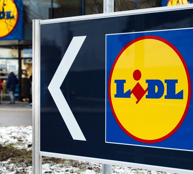  Das Bild zeigt ein großes Schild mit einem Pfeil und dem Logo des Supermarkts Lidl. Der Pfeil weist nach links, was darauf hinweist, dass die Filiale in diese Richtung liegt. Im Hintergrund sieht man den Eingangsbereich eines Lidl-Marktes mit einigen Personen, die sich in der Nähe befinden. Der Boden ist teilweise mit Schnee bedeckt, was auf winterliche Wetterbedingungen schließen lässt. | © AdobeStock/fotofotofoto