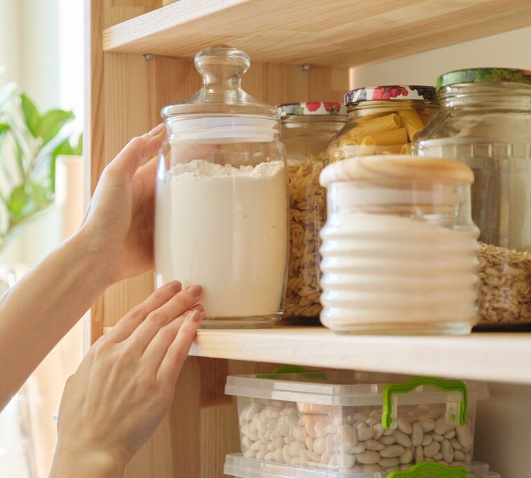 Eine Hand greift in einer hellen Küche nach einem großen Glas mit Mehl, das in einem Holzregal zwischen weiteren Vorratsgläsern steht. | © Adobe Stock / Valerii Honcharuk