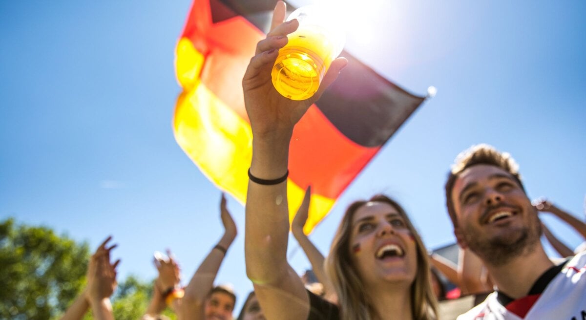 Jubelnde junge Fußballfans stoßen mit Bier an, schwenken eine Deutschlandflagge und feiern unter strahlend blauem Himmel.
