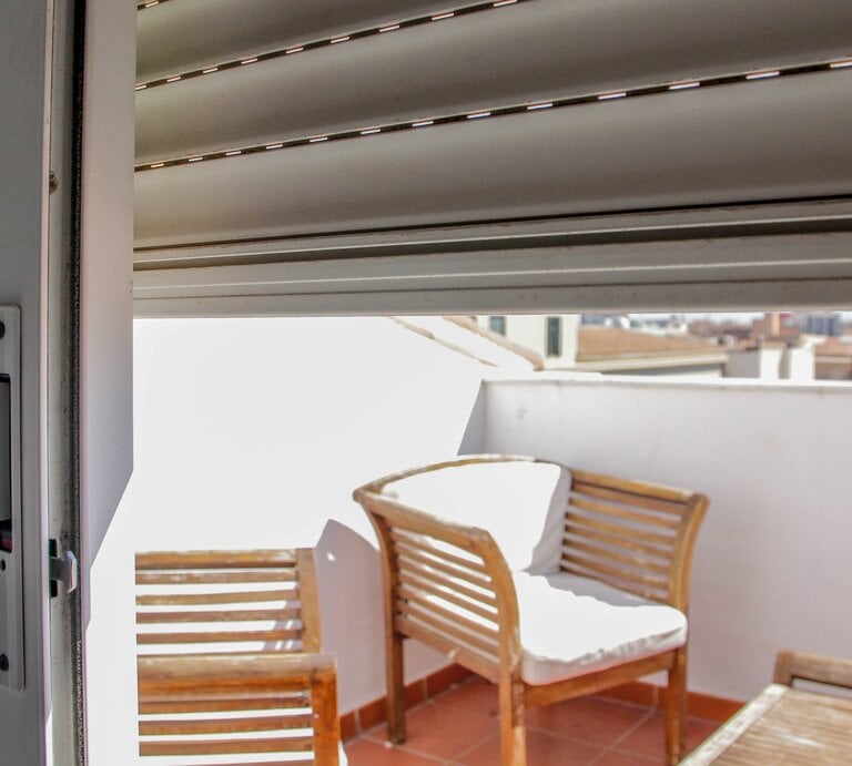 Ein Rollo ist halb heruntergelassen, eine Balkontür ist geöffnet und gibt den Blick auf eine Terrasse mit einem Tisch und einem Stuhl aus Holz mit weißem Bezug frei. | © gettyimages/ Kinga Krzeminska