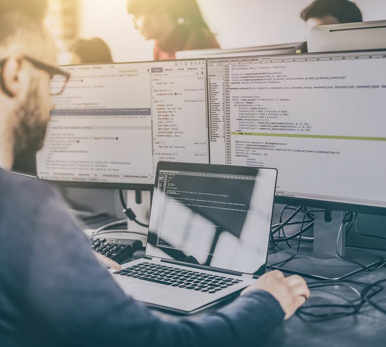 Zu sehen ist ein junger Mann der vor einem Laptop und zwei Bildschirmen sitzt. | © Shutterstock/ REDPIXEL.PL