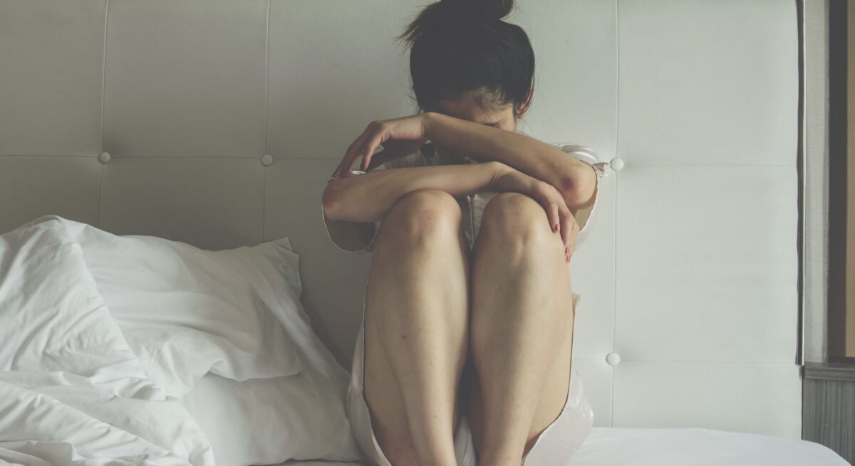 Junge Frau in hellem Nachthemd sitzt verzweifelt mit angezogenen Knien auf einem ungemachten Bett.