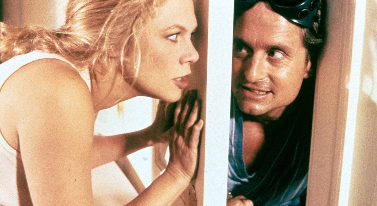 Eine blonde Frau im weißen Top lehnt sich angespannt an eine geöffnete Tür, durch deren Spalt ein Mann in blauem Hemd schaut.