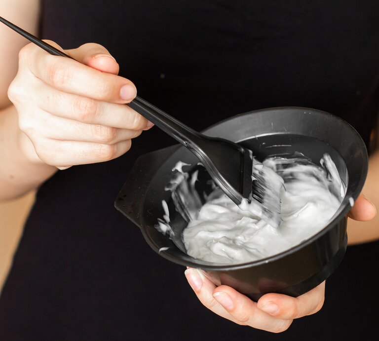 Hände einer Frau halten eine schwarze Schale mit cremiger Haarfarbe, während sie die Farbe mit einem schwarzen Pinsel rührt. | © Adobe Stock/ vladim_ka