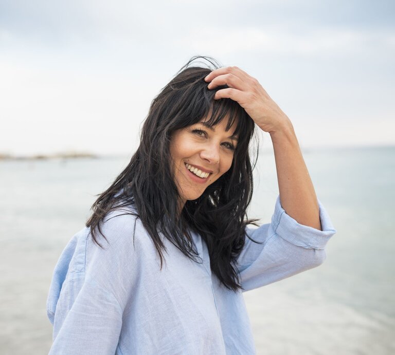 Lachende dunkelhaarige Frau mittleren Alters in hellblauer Bluse streicht sich am Strand fröhlich das Haar aus dem Gesicht. | © Getty Images/ Westend61