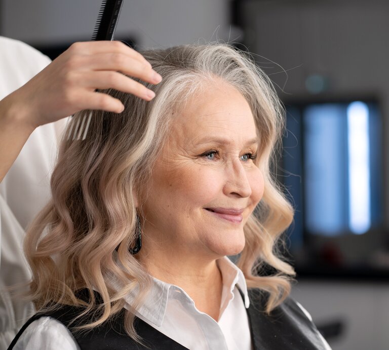 Eine Hand im weißen Ärmel kämmt weiche, blonde Locken; im Hintergrund ein dunkel eingerichteter Friseursalon. | © Adobe Stock/ Анжелла Антоновская