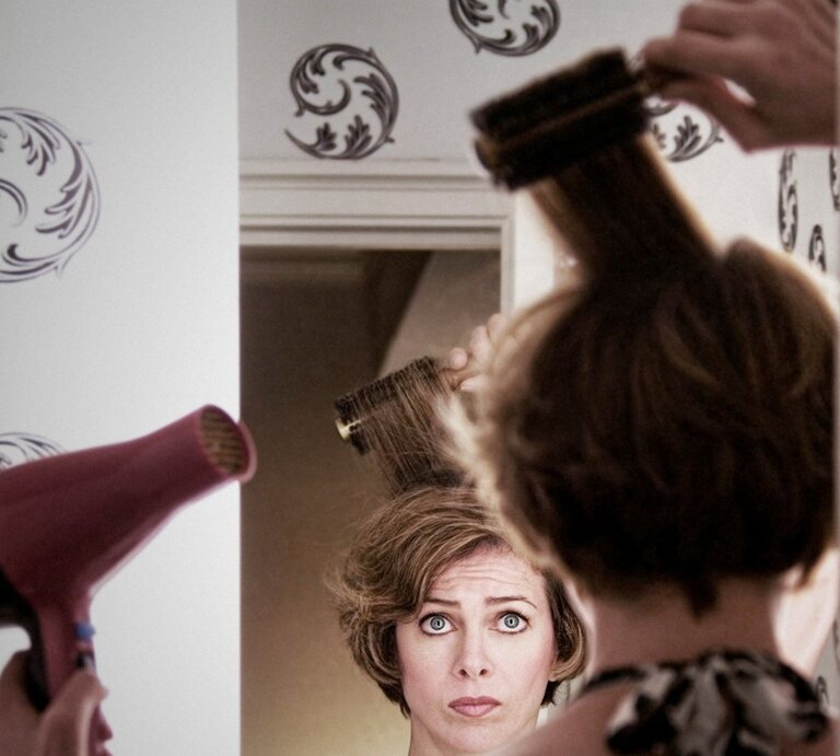 Eine Frau mit kurzen braunen Haaren schaut ernst in den Spiegel, während sie mit Rundbürste und Föhn ihre Haare stylt. | © Getty Images / Christopher Winton-Stahle