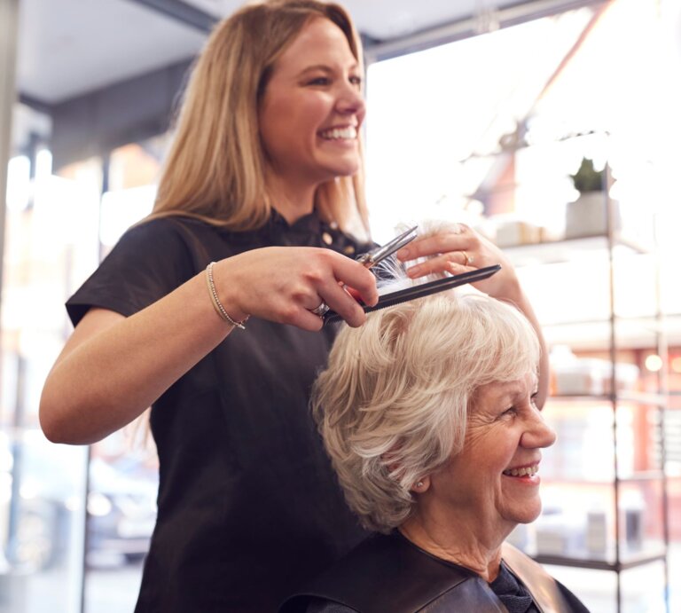 Eine junge Friseurin mit langen blonden Haaren schneidet lächelnd die Haare einer fröhlichen älteren Dame in einem hellen Salon. | © Adobe Stock/ Monkey Business