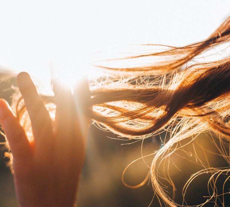 Nahaufnahme einer Hand, die durch langes Haar gleitet, das in der warmen Abendsonne leuchtet und sich im Wind bewegt. | © Adobe Stock/ KoralFox