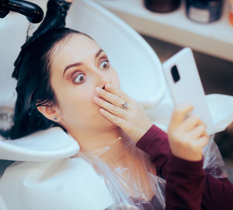 Junge Frau mit dunklem Haar hält schockiert das Handy hoch, während ihr beim Friseur die Haare gewaschen werden. | © Adobe Stock / nicoletaionescu