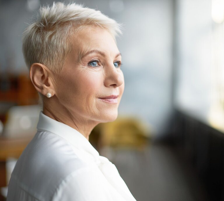 Reife Frau mit stylischer Pixie-Frisur - einer Kurzhaarfrisur für dünnes Haar - und Perlenohrringen trägt weiße Bluse und schaut verträumt ins Tageslicht. | © Adobe Stock/ shurkin_son