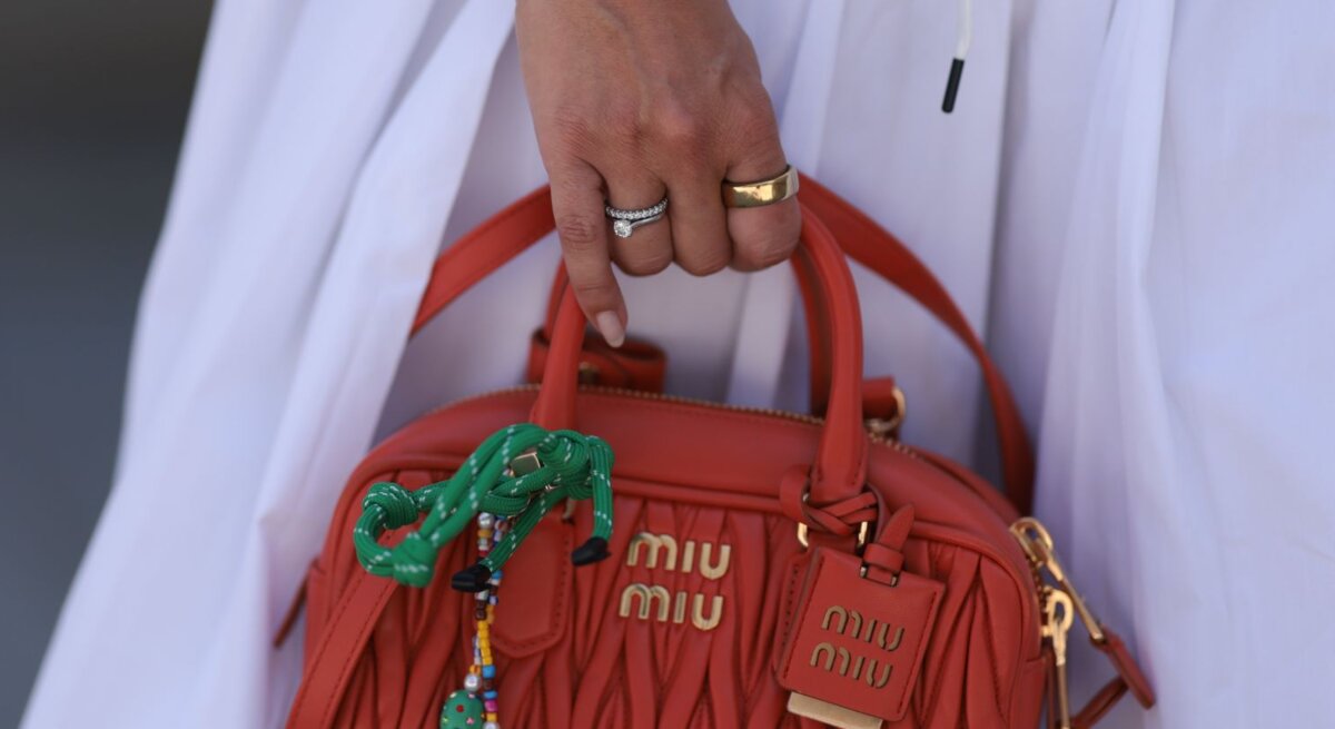 Hand mit goldenen und bunten Armbändern hält eine kleine orangene Miu Miu-Tasche mit dekorativen Elementen, vor einem weißen Kleid.