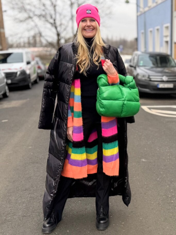Renate Zott mit pinker Mütze, langem schwarzen Steppmantel und bunt gestreiftem Schal steht mit grüner Tasche auf einer Stadtstraße. | © Renate Zott