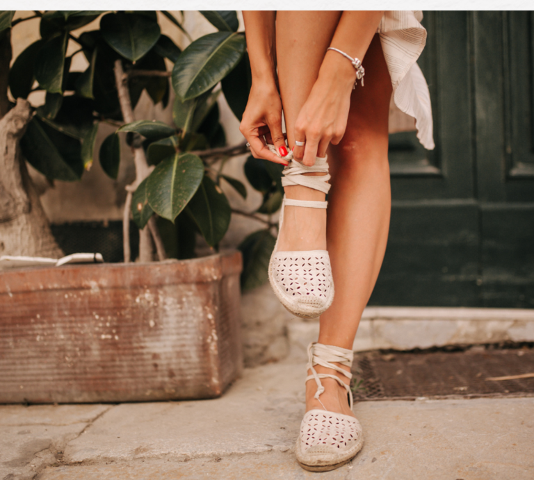 Zwei beige Wildleder-Sandalen mit goldenen Schnallen liegen auf zerknittertem, weißem Stoff im warmen Sonnenlicht. | © Canva/Collage:bildderfrau.de