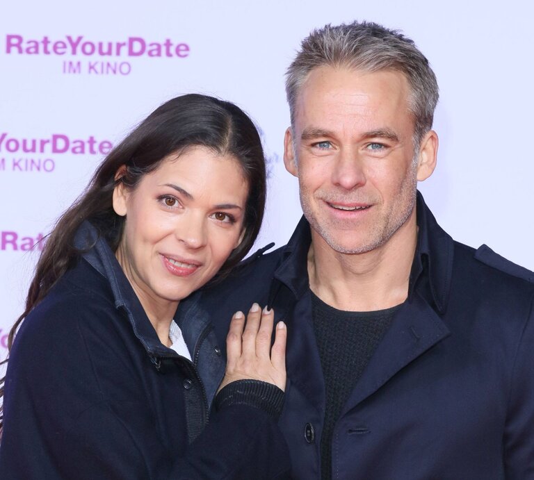 Katja Woywood und Marco Girnth  lächeln und posieren gemeinsam für ein Foto. Katja Woywood lehnt sich an Marco Girnth und schaut direkt in die Kamera, während Marco Girnth zur Seite blickt. Beide tragen dunkle Oberbekleidung. Im Hintergrund ist eine Werbetafel mit dem Schriftzug "RateYourDate IM KINO" in Pink zu sehen, was auf eine Filmpremiere oder ein ähnliches Event hinweist, bei dem sie anwesend sind. | © IMAGO / Tinkeres