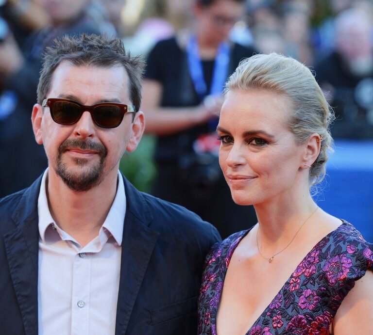 Alex Silva und Nina Hoss stehen Seite an Seite und blicken in die Kamera. Alex Silva trägt eine schwarze Jacke und Sonnenbrille, während Nina Hoss ein Kleid mit Blumenmuster und ein dezentes Make-up trägt. Ihr Haar ist zu einem eleganten Knoten gesteckt. Sie scheinen auf einem roten Teppich zu stehen, erkennbar an den unscharfen Gestalten und Kameras im Hintergrund, was auf eine Film- oder Unterhaltungsveranstaltung hindeutet. | © IMAGO / Manfred Segerer