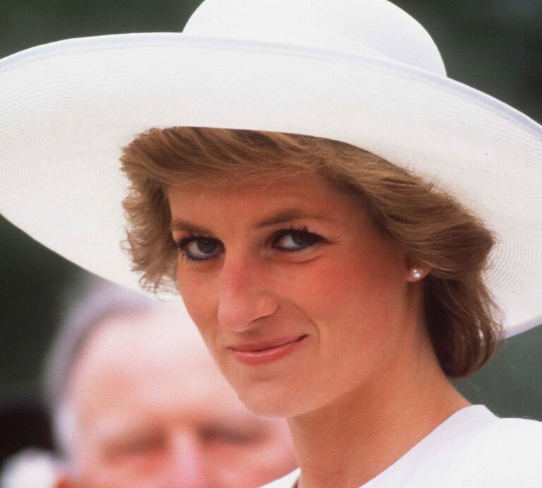 Das Bild zeigt Prinzessin Diana im Jahr 1989 mit ihrem charakteristischen kurzen Haarschnitt, die einen weißen Hut mit breiter Krempe trägt. Sie hat ein freundliches Lächeln und trägt schlichten Schmuck. Prinzessin Diana blickt sanft zur Seite und trägt ein Outfit, das Eleganz und Raffinesse ausstrahlt. Das Bild fängt einen Moment der Ruhe und des Selbstbewusstseins ein, mit einem unscharfen Hintergrund, der die Aufmerksamkeit auf Prinzessin Diana im Vordergrund lenkt. | © IMAGO / ZUMA Wire