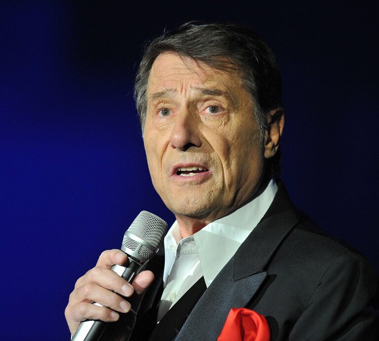 Udo Jürgens trägt einen dunklen Anzug mit rotem Einstecktuch und spricht oder singt in ein Mikrofon vor einem blauen Hintergrund. | © IMAGO / Revierfoto