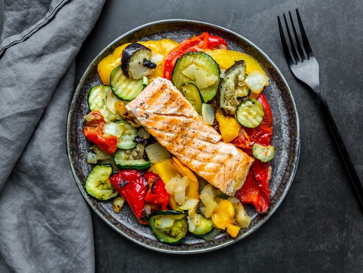 Das Bild zeigt einen Teller mit einer gesunden Mahlzeit, bestehend aus einem Stück gegrilltem Lachs auf einem Bett aus verschiedenen gegrillten oder gebratenen Gemüsen. Die Gemüsesorten scheinen Zucchini, Paprika und Zwiebeln zu umfassen