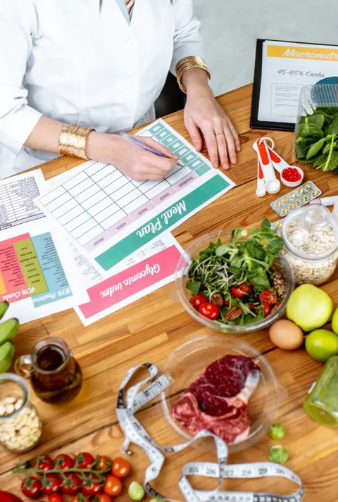Ernährungsberaterin erstellt einen Ernährungsplan am Tisch, umgeben von frischen Lebensmitteln wie Obst, Gemüse, Fleisch, Nüssen und Vollkornprodukten sowie Unterlagen zu Makronährstoffen und einem Maßband.