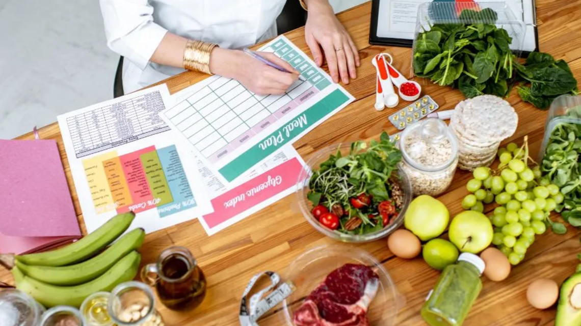 Ernährungsberaterin erstellt einen Ernährungsplan am Tisch, umgeben von frischen Lebensmitteln wie Obst, Gemüse, Fleisch, Nüssen und Vollkornprodukten sowie Unterlagen zu Makronährstoffen und einem Maßband.