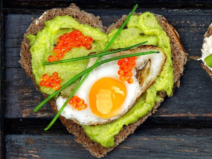 eiweiss-diaet.webp Herzförmiges Vollkornbrot mit Avocado-Creme, Spiegelei, rotem Kaviar und Schnittlauch auf dunkler Holzunterlage – gesundes, eiweißreiches Frühstück.