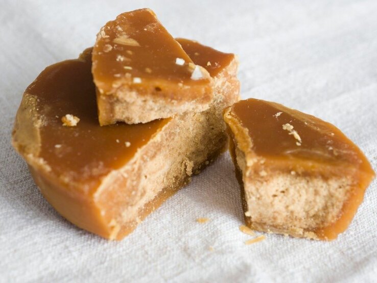 Das Bild zeigt Stücke von Jaggery, einem traditionellen, unraffinierten Zuckerprodukt, das oft in Südasien verwendet wird. Die goldbraune Färbung und die erkennbare Körnung weisen auf die natürliche Beschaffenheit hin. Der Jaggery scheint frisch gebrochen zu sein, was durch die unregelmäßigen Kanten und die sichtbaren Kristalle innerhalb des Zuckers deutlich wird. Die Textur sieht klebrig und feucht aus, typisch für hochwertigen Jaggery. Er liegt auf einer weißen Unterlage, die seine rustikale, natürliche Ästhetik betont.