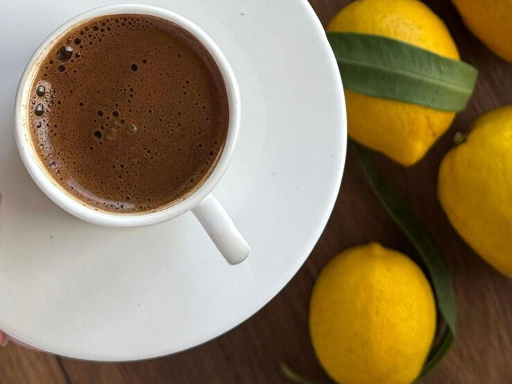 Tasse mit frisch aufgebrühtem, schaumigem Kaffee auf einer weißen Untertasse, daneben mehrere gelbe Zitronen auf einer Holzoberfläche.