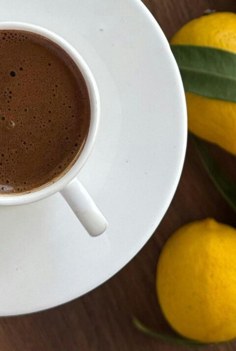 Tasse mit frisch aufgebrühtem, schaumigem Kaffee auf einer weißen Untertasse, daneben mehrere gelbe Zitronen auf einer Holzoberfläche.