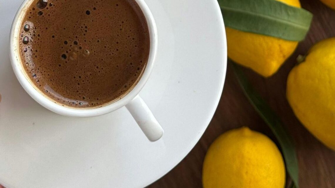 Tasse mit frisch aufgebrühtem, schaumigem Kaffee auf einer weißen Untertasse, daneben mehrere gelbe Zitronen auf einer Holzoberfläche.