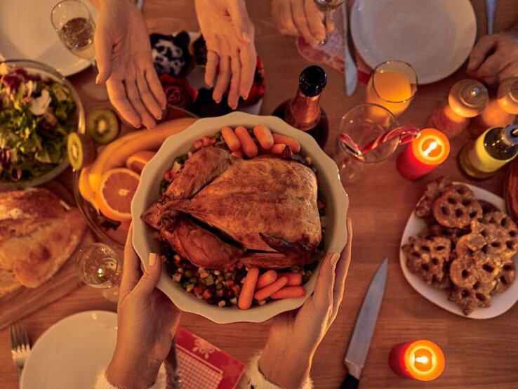 Hände reichen eine Schüssel mit gebratenem Geflügel und Gemüse über einen festlich gedeckten Tisch mit Kerzen, Getränken und Weihnachtsgebäck.