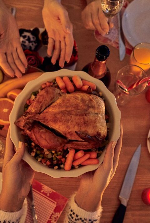 Hände reichen eine Schüssel mit gebratenem Geflügel und Gemüse über einen festlich gedeckten Tisch mit Kerzen, Getränken und Weihnachtsgebäck.