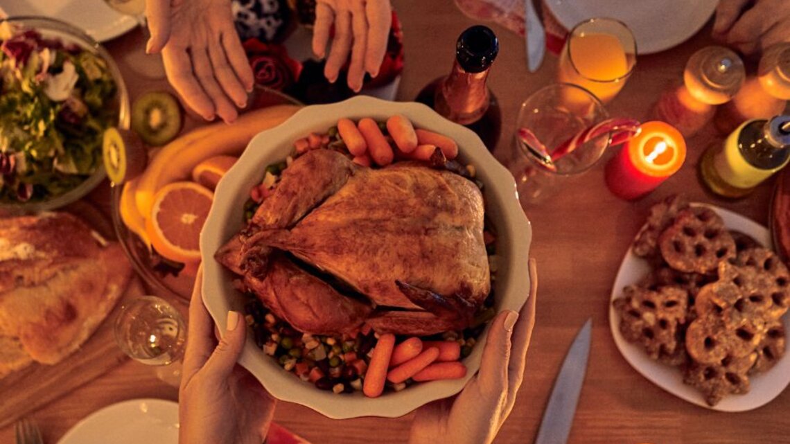 Hände reichen eine Schüssel mit gebratenem Geflügel und Gemüse über einen festlich gedeckten Tisch mit Kerzen, Getränken und Weihnachtsgebäck.