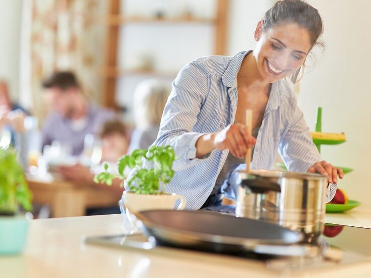 Eine lächelnde Frau in einem blau-weiß gestreiften Hemd steht an einem Herd und rührt mit einem Holzlöffel in einem Topf. Dampf steigt auf. Im Hintergrund sitzt eine Gruppe von Menschen an einem Tisch. Auf der Arbeitsplatte stehen eine Topfpflanze, eine Bratpfanne und eine Etagere mit Obst.