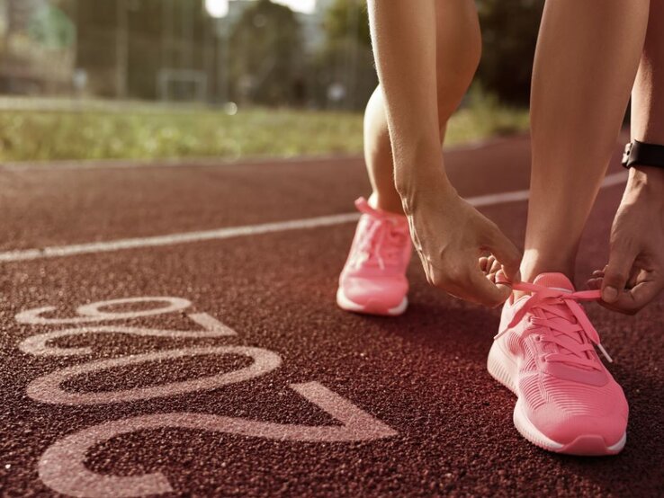 Eine Person bindet die Schnürsenkel ihrer pinken Sportschuhe, während sie auf einer Laufbahn steht. Im Vordergrund ist die Zahl "2026" auf dem Boden der Laufbahn zu sehen. Im Hintergrund sind unscharf Grünflächen und Gebäude erkennbar.