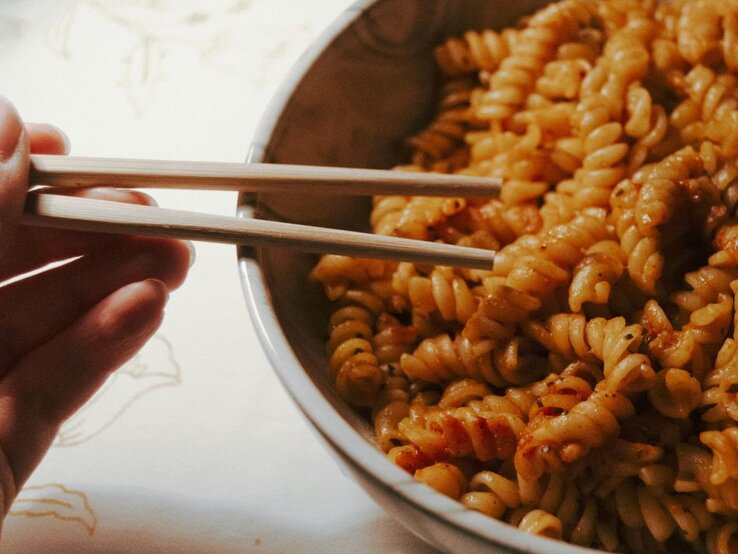 Hand hält Holzstäbchen über einer Schüssel mit Fusilli-Nudeln in rötlicher Soße auf hellem Untergrund.