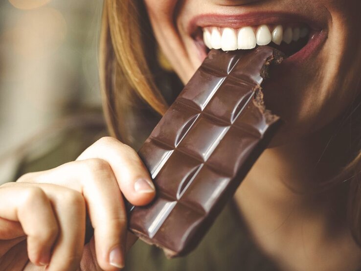 Eine Frau beißt genüsslich in eine Tafel dunkle Schokolade. Die Aufnahme zeigt den Moment des Genusses mit Fokus auf die Schokolade und die lächelnde Person.