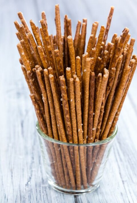 Ein Glas voller dünner, knuspriger Salzstangen, die senkrecht stehen. Das Glas steht auf einer hellen Holzoberfläche, der Hintergrund ist leicht verschwommen.