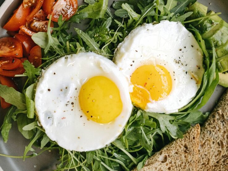 Zwei Spiegeleier mit leuchtend gelbem Eigelb, angerichtet auf frischem Rucola. Dazu gibt es frische, saftige Tomatenstücke, Avocadoscheiben und eine Scheibe Vollkornbrot, serviert auf einem grauen Teller.