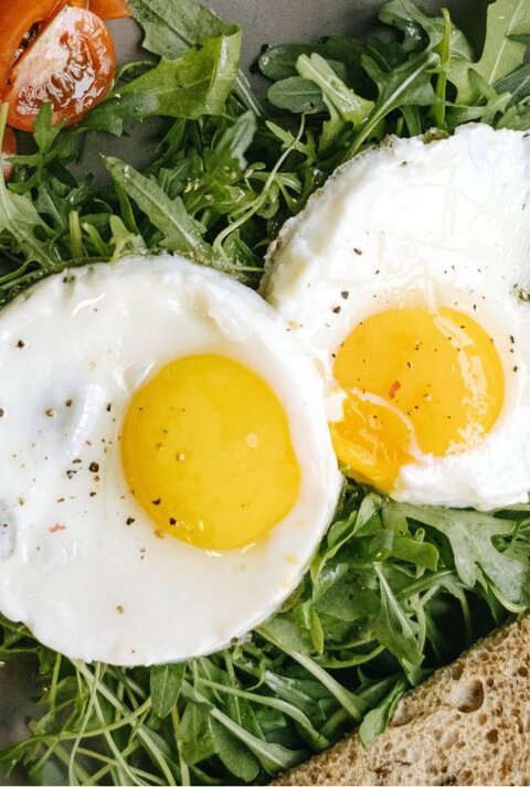 Zwei Spiegeleier mit leuchtend gelbem Eigelb, angerichtet auf frischem Rucola. Dazu gibt es frische, saftige Tomatenstücke, Avocadoscheiben und eine Scheibe Vollkornbrot, serviert auf einem grauen Teller.