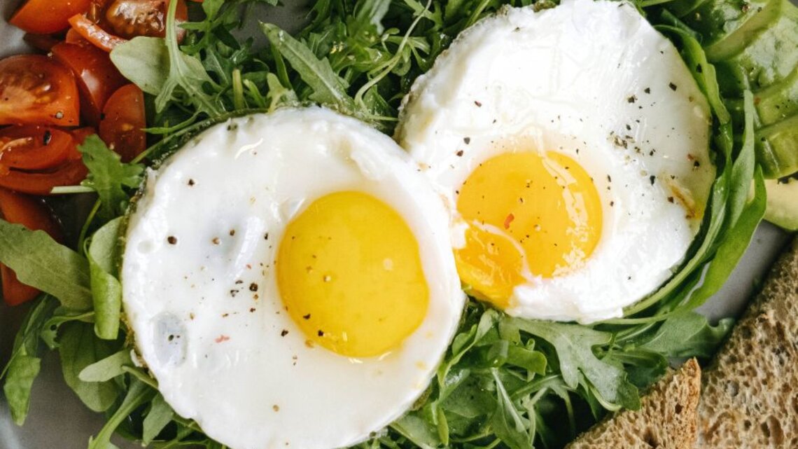 Zwei Spiegeleier mit leuchtend gelbem Eigelb, angerichtet auf frischem Rucola. Dazu gibt es frische, saftige Tomatenstücke, Avocadoscheiben und eine Scheibe Vollkornbrot, serviert auf einem grauen Teller.