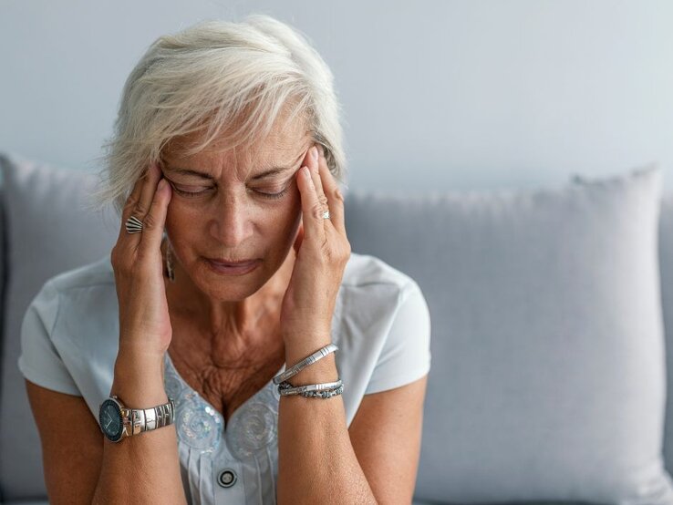 Frau mit grauen Haaren sitzt auf einem Sofa und hält sich mit beiden Händen die Schläfen.