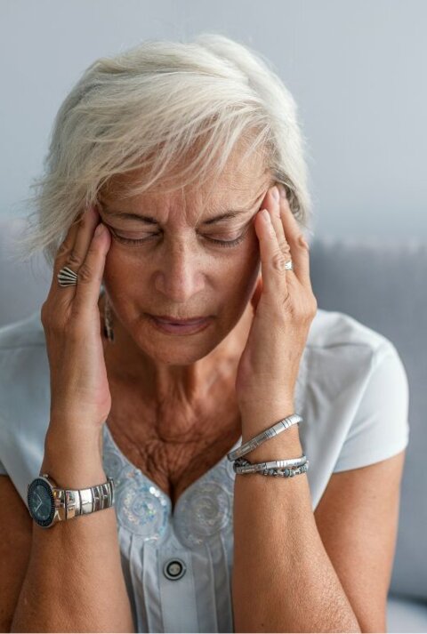 Frau mit grauen Haaren sitzt auf einem Sofa und hält sich mit beiden Händen die Schläfen.